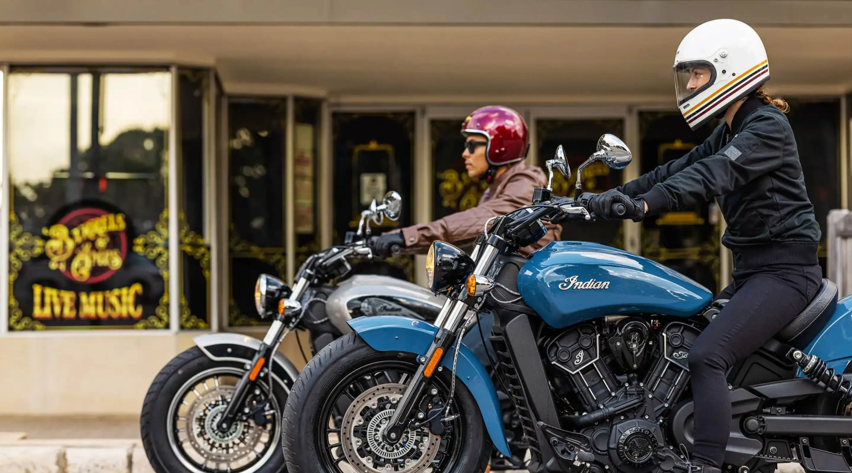 Two motorcyclist on Indian Motorcycles in front of slightly blurred building in the day time